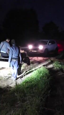 The video begins with a man in a gray sweatshirt and dark pants standing facing away from the camera, his left arm raised. To his right, a man in a blue jacket and dark pants is walking away from the camera, his back to the viewer. He appears to be carrying something green in his right hand. Behind him, a white SUV with its headlights on is partially visible, its rear red taillights illuminated. The vehicle is on a dirt or gravel road. To the left of the frame, a dark-colored car, possibly a sedan, is parked. Grass and foliage are visible on the right and left sides of the road, and trees are in the background against a dark night sky. The scene suggests a nighttime outdoor setting, possibly a rural or unpaved area. The overall impression is one of activity or an unfolding event.