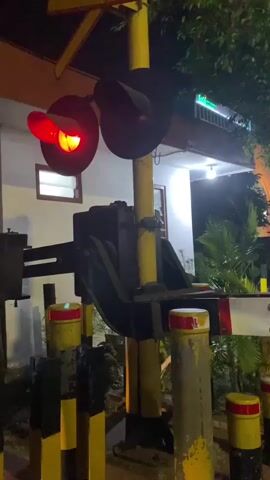 The video shows a railway crossing signal at night. The signal consists of two red lights that are illuminated, indicating that a train is approaching or present. The signal pole is yellow and black. The background shows a building with some lights on and some greenery. A white barrier arm is visible, suggesting the crossing is about to close. There are no people visible in the video.