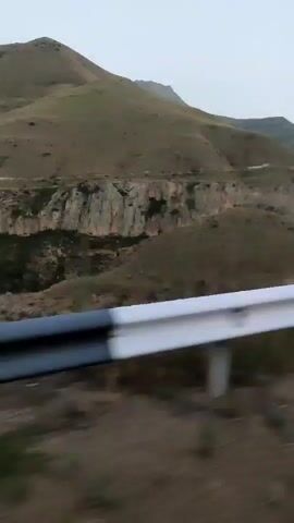 The video shows a landscape of rolling hills and mountains. A guardrail is visible in the foreground. The sky is overcast. The terrain is arid with sparse vegetation. The road appears to be winding through a canyon or valley.