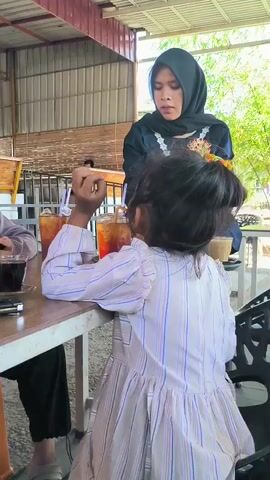 A young girl wearing a white striped dress is seated at a table in what appears to be an open-air restaurant or cafe. A woman wearing a black hijab is serving drinks, placing a glass of iced tea in front of the girl. Another person, presumably a teenager, is seated at the same table. The table also holds a small cup of coffee and an iced drink. The setting looks somewhat casual with a metal roof structure and glimpses of trees outside.