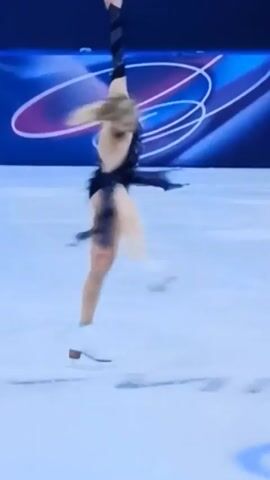 The video shows a woman figure skater in a black sparkly dress performing a back stretch move on a light blue ice surface.  She's blond with her hair pulled back.  She has her left arm up over her head and her right leg extended up in the air. The background is blue with some white designs.