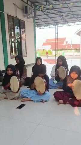 A group of girls, most of whom are wearing headscarves, are sitting on the floor outside a building and playing drums. The building has a green wall and a white awning. The girls are holding round, wooden drums with light-colored skins. Some of the girls are actively playing their drums, while others appear to be waiting their turn. In the background, there is a street with some buildings and trees. The overall atmosphere seems to be one of learning and practice.
