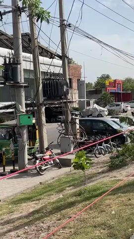 The video shows a street scene in Pakistan on a sunny day. The camera pans from a utility pole with electrical wires and equipment to a row of parked cars and vans along the side of the road. A Honda dealership is visible in the background. The camera then moves onto the road, following a motorcycle rider. Other motorcycles and cars are on the road. Street signs for "Kashmir Road" and "Gattwala" are visible. Trees line the side of the road. The sky is blue with scattered clouds.