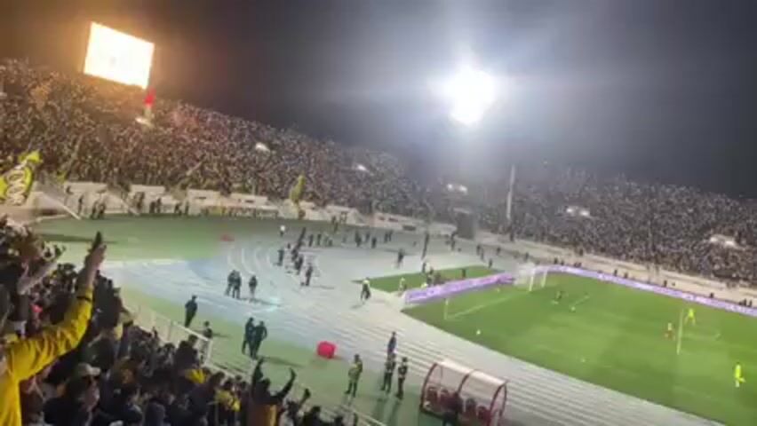 The video shows a soccer game at night in a packed stadium. The stands are full of people, mostly dressed in white, with some sections wearing yellow. A bright stadium light illuminates the field where players in yellow and blue uniforms are visible. Security personnel stand along the track surrounding the field, facing towards the stands. A few individuals are walking on the track. The video is filmed from an elevated position behind a team bench, where fans in yellow are cheering and raising their arms. A scoreboard is visible in the background, displaying a digital graphic.