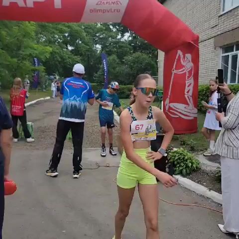 A young female athlete wearing a sports bra and neon yellow shorts with bib number 67 crosses an inflatable archway. She is wearing large, dark sunglasses and has her hair pulled back. She runs towards the camera and then turns and hugs another young woman wearing an orange athletic top and skirt. The second woman is wearing a white headband and has her dark hair in a braid. They speak to each other briefly. A woman wearing a white headscarf and a striped jacket stands in the background. Another person, partially visible, stands behind her. A man wearing a blue shirt with a mountain design and dark pants is visible on the left side of the screen. Behind the archway, a young male athlete in a blue shirt and cap is visible. The scene appears to be an outdoor sporting event with a building and greenery in the background.