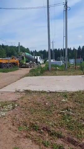 The video captures an active construction site on a clear, sunny day in what appears to be a rural or semi-rural setting. A prominent yellow concrete pump truck, identified with "ЗОРКОВ" (ZORKOV) and "КОМПАНИЯ" (COMPANY) text, along with a phone number "+7(495)333-33-33," is seen extending its long, articulated boom over an incomplete building structure made of cinder blocks. Construction workers are visible on top of the building, guiding the concrete as it's poured.

A green and white concrete mixer truck, bearing the word "БЕТОН" (BETON) on its side, is positioned behind the concrete pump truck, supplying the material. A dark gray Nissan Pathfinder SUV is parked on the dirt ground next to the construction area. The foreground features a dirt road with scattered patches of grass and some concrete slabs. Utility poles with overhead power lines traverse the scene, and a dense forest lines the horizon under a bright blue sky with some clouds. The activity clearly shows a concrete pouring operation, involving heavy machinery and manual labor. The person speaking in the video's audio notes the "heavy work" being performed.