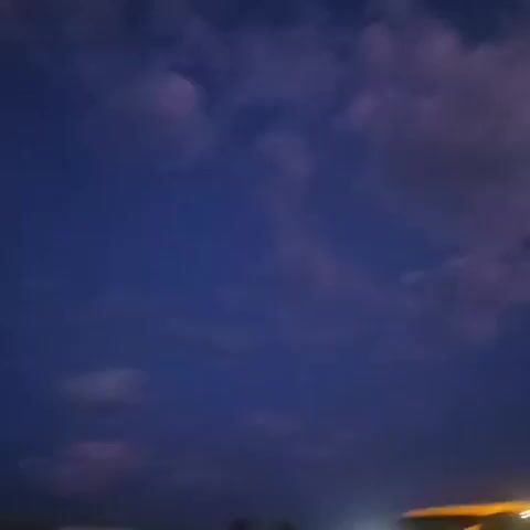 The video shows a street scene at dusk or night. In the first frame, a blue sky with purple clouds is visible, along with a street light and a blurry signpost. The subsequent frames show streetlights illuminating a road with cars, and the sky gradually darkens to a deep blue or purple. In the final frame, a crescent moon is visible in the dark sky. The overall mood is calm and somewhat melancholic, due to the low light and the twilight setting.