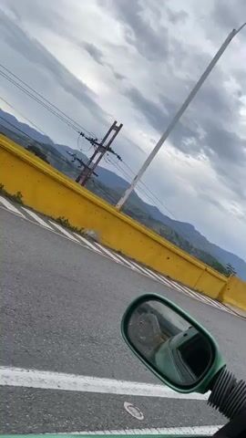 The video begins with a close-up of a car's side mirror, reflecting a cloudy sky and a mountainous landscape. The camera then pans to the right, revealing a road with a yellow barrier on the left and a row of streetlights stretching into the distance. Several cars are visible on the road ahead. The perspective suggests the video is being filmed from inside a moving vehicle, likely a car or motorcycle. The sky is filled with dramatic, grey clouds, hinting at possibly inclement weather or an impending storm. The overall scene depicts a journey along a scenic, elevated road, possibly a highway or bridge, with a backdrop of natural mountain terrain.
