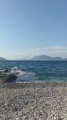 This media file captures a serene view of a pebble beach on a clear day. The foreground is dominated by a textured expanse of smooth, light-colored pebbles. Beyond the beach, the deep blue sea stretches out, with gentle waves lapping against a rocky shoreline. In the distance, a chain of hazy, blue-green mountains rises against the horizon, partially obscured by wisps of white cloud. The sky above is a vast, unblemished expanse of pale blue, suggesting a calm and pleasant weather condition. The scene evokes a sense of tranquility and natural beauty, characteristic of a Mediterranean coastline. The time of day appears to be morning or early afternoon, given the bright, even lighting. There is no visible text in this media file.