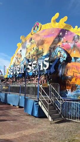 A colorful amusement park ride labeled "SEVEN SEAS" is visible from the side with a bright blue sky overhead. The ride is decorated with pirate-themed imagery, including ships and a pirate captain. People are seated on the ride, facing outwards. A set of metal stairs leads up to the ride entrance. The ground in front of the ride is a mix of dirt and gravel.