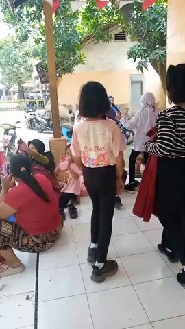 A group of people, mostly women and children, are gathered on a tiled outdoor patio. The children are playing and some adults are watching them. There are trees and buildings in the background. The atmosphere appears to be relaxed and casual.