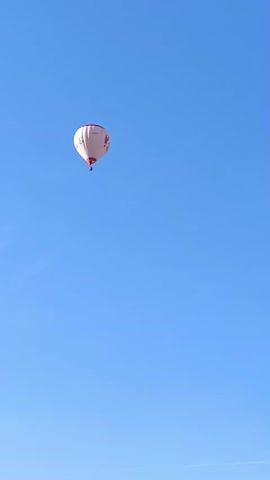 The video shows a hot air balloon floating in a clear blue sky. The balloon is white with red and black designs. It appears to be moving slowly, carried by the wind.  There is a faint contrail in the sky as well as the disembodied voice of a man.