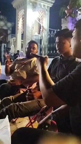 Several men are gathered outdoors at night, seemingly at a casual outdoor gathering or food stall. They are seated on folding chairs around a low table. In the foreground, a person's hand and arm are visible, holding something up, possibly a phone. To the left, a man with a beard wearing a light-colored t-shirt is gesturing with his hands, appearing to speak. Further to the left, another man in a light-colored polo shirt sits looking to his right. To the right of the gesturing man, a man in a dark t-shirt is looking to his left. Behind them, a white structure with arched openings, possibly part of a building or a decorative archway, is illuminated. In the background, a food stall with a lit sign and a water dispenser is visible, along with other people milling about. The overall atmosphere appears relaxed and social.