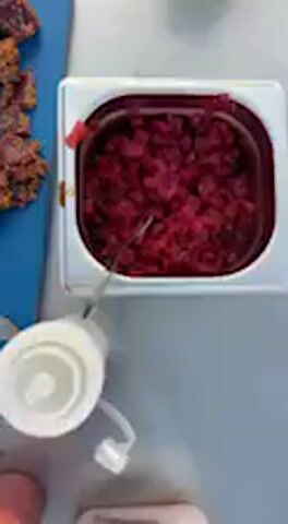 The video presents a top-down view of a food preparation area, likely in a kitchen or outdoor cooking setup in Barbate, Spain.

On a blue cutting board, several pieces of dark red, sliced meat, possibly tuna, are arranged next to a chef's knife. A small white bowl with a white granular substance, likely salt, is also visible.

Adjacent to the cutting board, a white rectangular container holds a substantial amount of finely chopped red food, which could be a relish, pickled onions, or beetroot salad. A spoon is inside this container. Another white rectangular container contains a brown, granular substance, possibly breadcrumbs or a spice mix.

A white box with the partial text "KON" is visible, with black gloves resting on it. A white rectangular plate displays several neatly arranged, longer slices of the dark red meat, possibly tuna, lightly seasoned. A small, round white plate holds a circular mound of finely diced red food, garnished with small, light-colored sprinkles, suggesting a tuna tartare or ceviche.

Hands are briefly visible in the frame, indicating active preparation. In the background, a person is heard speaking in Spanish, mentioning "una tosta de atún con una base de trufa" (a tuna toast with a truffle base) and describing "los tacos" (the pieces) as "fresquito" (fresh), confirming the presence of tuna and suggesting truffle as an ingredient. The scene captures the final stages of preparing and plating dishes.
