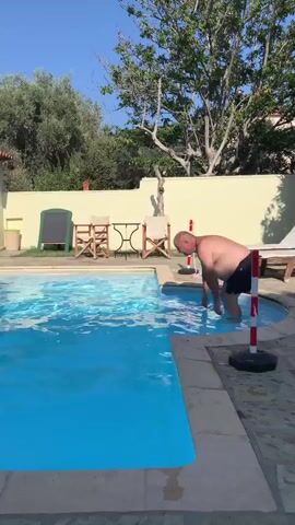 A man jumps into a swimming pool. He is wearing black shorts. The pool is blue and surrounded by a stone patio. Behind the pool is a yellow building with a red-tiled roof and an awning. There are trees and bushes in the background. The sky is clear and blue.