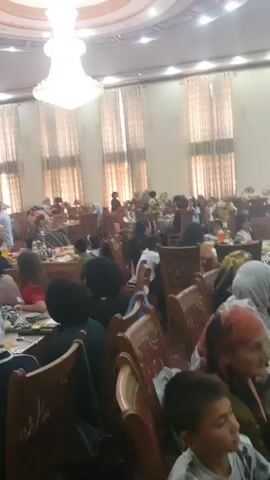 This video captures a lively indoor gathering, likely a celebration or a feast, held in Rizamahalla, Uzbekistan. The scene is set within a large, ornately decorated hall, illuminated by a grand chandelier hanging from the high, domed ceiling. Tall, cream-colored curtains adorn the numerous windows, adding to the elegant ambiance.

The hall is filled with people seated at round tables laden with food and drinks. The attendees, a mix of men, women, and children, appear to be engaged in conversation and enjoying the meal. Many women are wearing headscarves, typical of the region. The tables are set with an array of dishes, including bread, fruits like watermelon, and various small plates of food. Teapots and cups are also visible, suggesting that tea is being served.

In the background, a group of musicians can be seen, possibly providing entertainment for the event. A photographer with a camera is also present, documenting the occasion. The overall atmosphere seems joyful and communal. The lighting suggests it is likely daytime or a well-lit evening event. The décor and attire point towards a cultural celebration specific to Uzbekistan. No discernible text is visible in the video.