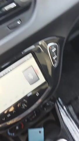 The video shows the interior of a car from the perspective of the passenger. The dashboard is visible in the foreground, with a blurred air vent. The view outside the passenger window is also visible, showing a green hedge and a building in the background. The car appears to be in motion.