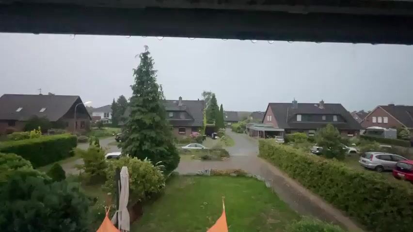 The video shows a view of a residential neighborhood in the rain. Several houses with dark roofs are visible, separated by trees and hedges. The sky is overcast and droplets cling to the edge of the window frame.  Cars are parked on the street and in driveways.  A lawn is visible in the foreground, with some orange fabric partially in view at the bottom of the frame. The rain is falling steadily.
