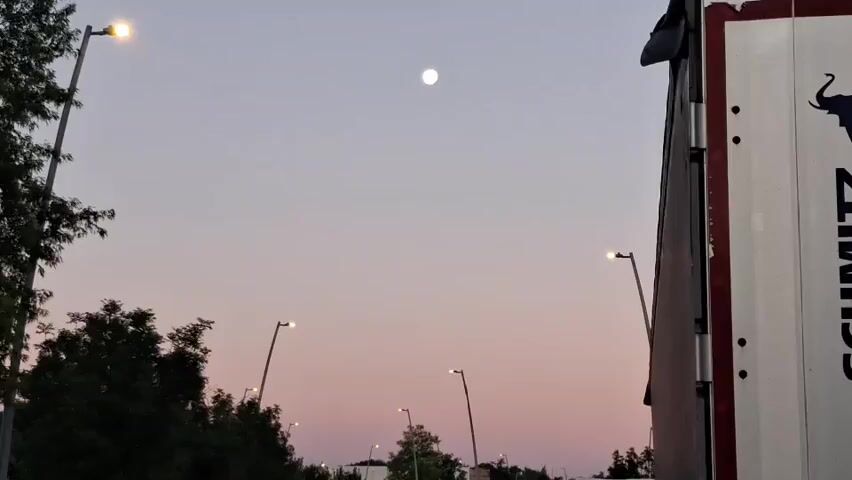 The video shows a twilight sky with the moon visible as a bright orb. Several streetlights are illuminated, casting a soft glow. On the right side of the frame, a portion of a large, white vehicle, possibly a truck or container, is visible with a dark logo or graphic on its side. Trees and other foliage are present on the left side of the frame, silhouetted against the lighter sky. The overall impression is of an urban or suburban setting during the transition from day to night.