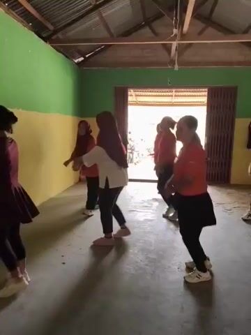 A group of at least six women are dancing in a room. The room appears to be a simple, enclosed space with a corrugated metal roof, wooden beams, and walls painted in green and yellow. There is an open doorway in the background leading to a brighter area. The women are dressed in casual clothing, with several wearing hijabs and red t-shirts. They are all facing towards the camera and appear to be engaged in synchronized movements. The floor is concrete and there are shadows cast on it, suggesting the presence of light sources. A fan is visible on the right side of the frame.