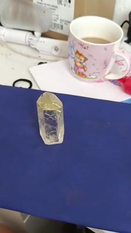 The video displays a clear, faceted crystal, likely quartz, positioned centrally on a textured blue surface. The crystal is elongated and hexagonal, with a pointed apex. Inside the crystal, there appear to be inclusions or internal fractures, creating a web-like pattern. The lighting suggests an indoor setting, possibly a desk or workspace, as indicated by the blurred background elements.

In the background, out of focus, a pink mug with a cartoon illustration of a fox and rabbit is visible. To the left of the mug, a white object, possibly an electronic device or a tool, is also blurred. A white sheet of paper lies beneath the mug. The overall scene is static, with no discernible activity or event taking place beyond the presentation of the crystal. The time of day and weather are not ascertainable from the visual information. No text is clearly legible in the video. The setting is not specifically identifiable as Nizhnekamenka, Russia, based on the provided image content.