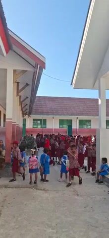 This is a vertical video of an outdoor school scene. In the foreground, several young students in uniforms are walking. Some are wearing red and white patterned shirts and red shorts or skirts, while others are in blue shirts and shorts. Many of them are carrying backpacks. In the background, a larger group of students, also in uniform, are gathered in front of a school building. The building has a red tiled roof and white and red exterior walls with green windows. The sky above is clear and blue. The lighting suggests it is daytime, likely morning or late afternoon.