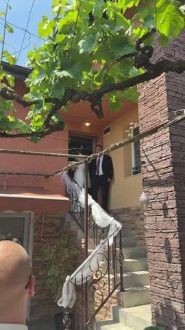 A video captures a couple on their wedding day. The bride wears a white wedding gown and veil, while the groom wears a black suit, white shirt, and purple tie. They stand at the top of a stone staircase adorned with white tulle, with green leaves from a tree overhead. A person in a white shirt and sunglasses is visible at the bottom of the frame, partially obscured. The house behind them is a reddish-brown color with a matching door. As the video progresses, the bride and groom descend the stairs, pausing to pose for photos before continuing down.
