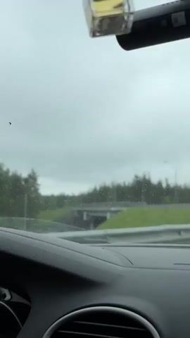 The video shows a car's dashboard and windshield view while driving on a highway on a cloudy, possibly rainy day. Trees are visible in the distance and the road ahead is wet. There is a bridge over the road in the distance. A dashcam is visible on the windshield. The music playing suggests someone is driving for pleasure.