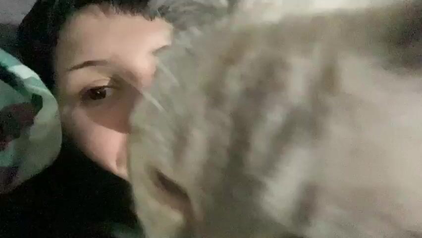 A close-up shot shows a person lying down with their face turned towards the camera. A light-colored cat is in the foreground, obscuring part of the person's face. The person has dark hair with bangs and visible piercings on their lip. The cat has light-colored fur and is looking directly at the camera. The background is dark and appears to be indoors, possibly in bed, with some blurry green fabric visible on the left. The lighting is dim.