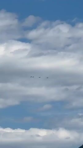 A vertical shot shows a cloudy blue sky. Three small airplanes are flying in a tight formation in the middle of the frame. The planes are dark silhouettes against the sky and clouds.