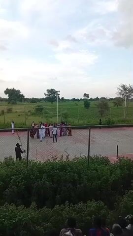 The video begins with a wide shot of an outdoor event. A group of people, predominantly women in colorful attire and men in white clothing, are gathered in a circle on a paved area surrounded by a fence. In the background, there is a large green field with trees and a cloudy sky. The camera then pans to a seating area under a roof where a crowd of spectators is gathered, facing away from the camera. The focus shifts to individuals in the crowd, showing close-ups of young men observing the event. The overall environment appears to be a community gathering or a sporting event.