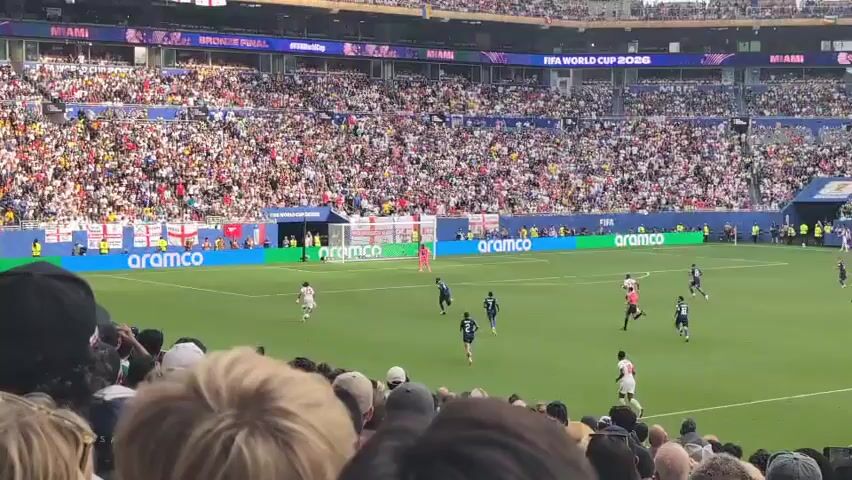 The video shows a soccer game taking place in a stadium filled with spectators. The field is green, with white lines marking the boundaries. Several players in white and blue uniforms are on the field, engaged in the game. A large crowd of people, dressed in various casual attire, are seated in the stands, many of them holding up their phones, likely to record the event. The stadium appears to be modern, with tiered seating and banners displaying advertisements and event information, including "FIFA WORLD CUP 2026" and "MIAMI." The sky is visible above the stadium structure.