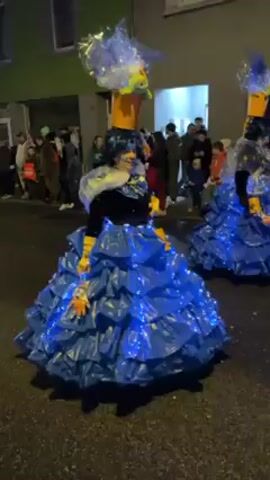 Two people are dressed in elaborate costumes made of blue plastic bags, illuminated by blue Christmas lights. They are participating in a parade or street performance. The costumes have a voluminous, gown-like shape and the performers wear orange hats, one of them decorated with a plume. Other people line the street, watching and recording with their phones. The setting appears to be a residential street at night, with buildings visible in the background. An orange clad person with a music player walks between the two performers.