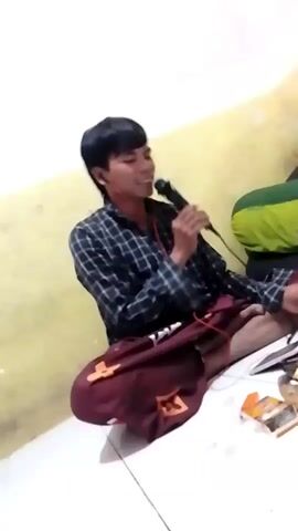 A young man with short, dark hair and a plaid shirt is sitting on the floor, holding a microphone and speaking into it. He is wearing headphones with red wires. He has on a maroon sarong with a pattern. He is in a room with a yellow wall and a white floor. There is a cigarette pack on the floor in front of him.