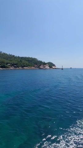 The video shows a view of a tropical island coastline from the water. The water is a clear turquoise blue. The island is lush with green vegetation, including palm trees. Along the shore are several small buildings. The sky is clear and blue.  The camera appears to be on a moving boat, evident from the slight movement and the wake in the water.