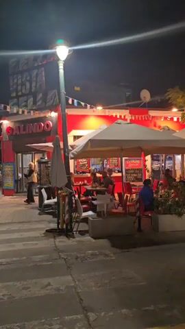 The video shows a street scene at night. The street is paved and appears to be a pedestrian zone or a street with limited traffic, with visible crosswalk markings. In the foreground, there is a restaurant named "Galindo" with a bright red facade. The restaurant has outdoor seating under white umbrellas. People are seated at the tables, and one person is standing near the entrance. The street is illuminated by lampposts and the lights from the buildings. Bunting decorations are strung across the street. A white car is seen driving across an intersection. Other buildings, trees, and street furniture are visible in the background. Overall, the scene suggests a lively nighttime atmosphere in a commercial area.