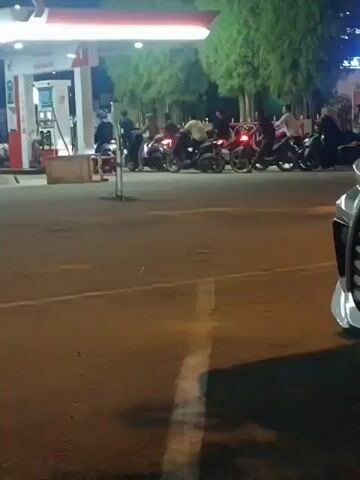 The video depicts a nighttime scene at a gas station. Several motorcycles are parked in a line, with people sitting on or standing near them. A gas pump with its canopy is visible on the left side of the frame. Trees are present behind the motorcycles. The ground is paved, and a white line marks a lane on the road in the foreground. Part of a car is visible on the right side of the frame, obscuring some of the scene. The lighting suggests it is dark, with artificial lights from the gas station and possibly surrounding areas illuminating the scene.