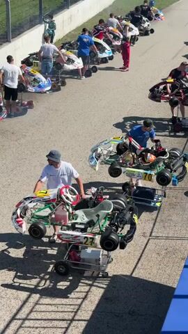 The video shows a group of people and go-karts on a paved surface, likely a racetrack. Several go-karts are lined up, some with people standing next to them. One person in the foreground is adjusting a go-kart. Another person in the background is wearing a red racing suit. The environment appears to be outdoors on a sunny day, with shadows visible on the ground.