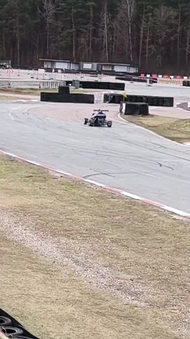 The video shows a go-kart driving on a gray racetrack. The go-kart is dark in color and has a visible driver. There are tire barriers along the sides of the track. A person is standing next to a tire barrier on the side of the track, observing the go-kart. The track is surrounded by grass and a forest in the background. There are some buildings and other go-karts in the distance. Tire tracks are visible on the gray track surface. The sky appears overcast.