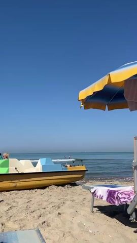 A sunny day at the beach. A yellow pedal boat with blue and green seating is pulled up on the sandy shore. In the background, the calm, blue ocean meets the clear sky. Two people are visible in the water, wading or swimming. A striped beach umbrella, yellow and blue, is open and angled, casting a shadow. A lounge chair with a purple and white towel draped over it is positioned in the foreground, to the right of the pedal boat. The sand appears dry and light-colored.