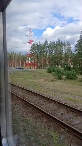 The video shows a view from a moving train. Twin railway tracks run parallel to the camera's perspective, heading towards the horizon. To the right of the tracks, a grassy embankment slopes upwards towards a dense forest of pine trees. A large, red and white radio tower stands prominent within the treeline, with a clear blue sky and white clouds overhead. A few cars are visible on a road in the distance, to the left of the radio tower. The camera is positioned to capture the passing scenery, emphasizing the motion of the train and the rural landscape.