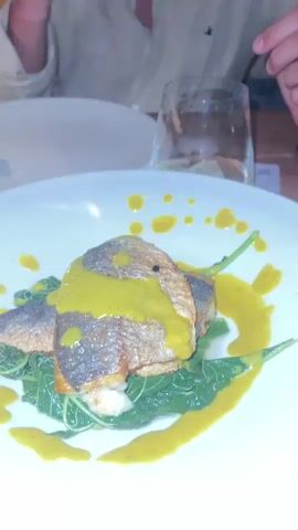 The video begins with a close-up shot of a plate of food, featuring what appears to be pan-seared fish with a yellow sauce and greens. The camera then pans to a man sitting at a table, smiling and looking directly at the camera. He is wearing a beige linen shirt. The scene appears to be a restaurant at night, with other diners visible in the background. The video continues to show more food on plates, including what looks like a seafood risotto, and hands reaching for food. There are glasses of white wine on the table. The lighting is dim, suggesting an evening setting.