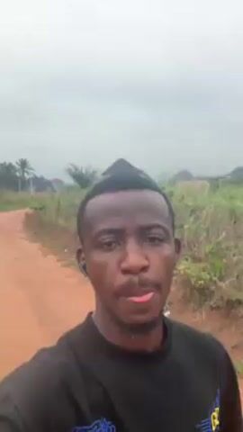A man records a selfie video outdoors on a dirt path. He is wearing a black t-shirt and has earbuds in. There are houses and trees visible in the background, along with vegetation on either side of the path. The sky is overcast. The man is looking directly at the camera and talking.