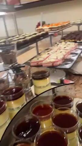 The video shows a dessert buffet on a countertop in a room. There are several different types of cakes, pastries, and puddings on display. The desserts are arranged on trays and plates, with tongs for serving. There is a stack of glasses nearby. The countertop is made of a light brown, speckled material. The room has light-colored walls and a wood-paneled section below the countertop.