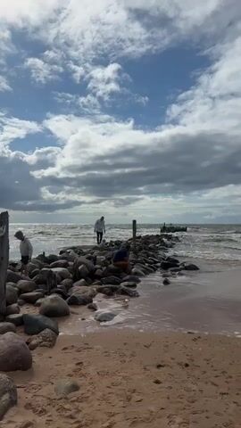 A video shows a sandy beach with a breakwater made of large rocks extending into the ocean. Three people are visible on the breakwater. The person on the left is a child wearing a grey hoodie and dark shorts, standing on the rocks. In the middle, a person in a white top and dark pants is walking towards the end of the breakwater. To the right, a person in a dark blue t-shirt and dark shorts is crouched down on the rocks, appearing to be examining something. The ocean has moderate waves, and the sky is partly cloudy with blue patches. The overall scene depicts a somewhat overcast but active day at the beach.