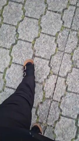 The video begins with a close-up, blurred shot of a person's legs and feet walking on a cobblestone-like pathway. The person is wearing dark pants and tan shoes. The view then shifts to show a residential street in Evere, Belgium. A multi-story brick building is under renovation, indicated by scaffolding and green mesh covering parts of the facade. A tall ladder leans against the building, and construction materials like wooden pallets and what appears to be roofing tiles are visible near the entrance. Several cars are parked along the street in front of the building. The sky is overcast, suggesting a cloudy day. The camera continues to pan and tilt, offering brief glimpses of the street and the building from different angles, interspersed with more shots of the cobblestone pathway. The overall impression is a candid, possibly personal, recording of a walk through a residential area undergoing some construction or repair work. The time of day appears to be late afternoon or early evening, given the lighting.