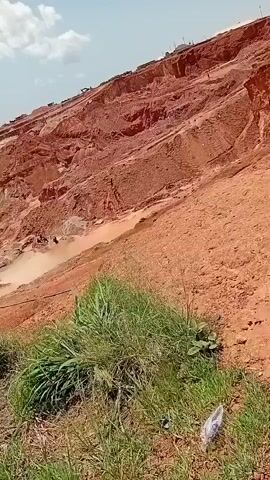 The video shows a hillside with red soil. There are several people working on the hillside. Some people appear to be digging or excavating the soil, while others are carrying loads of soil. There are also some large vehicles, possibly excavators or trucks, visible on the upper part of the hillside. The environment appears to be arid, with sparse vegetation in the foreground. The sky is visible in the background and appears to be partly cloudy. The overall impression is of an active excavation or mining operation.