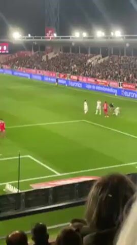 The video shows a soccer game from the stands. A crowd of spectators fills the stadium seating. On the field, players wearing red and white uniforms stand near the penalty spot. A player in a white uniform is kneeling. The score is tied 0-0. There are advertising signs along the field's edge. The field is green and brightly lit by overhead lights.