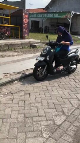 A woman wearing a hijab and a blue shirt is sitting on a black scooter. She is holding onto the handlebars and appears to be looking down. The scooter is parked on a paved area with patterned bricks. In the background, there is a building with a sign that reads "JUAL SAYUR SEGAR," indicating a fresh vegetable shop. To the left, there is a yellow awning and a food cart. The scene appears to be outdoors on a sunny day.