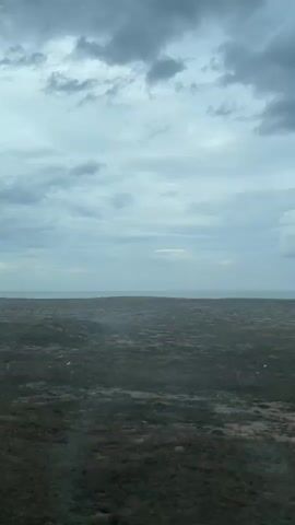 The video shows a wide, flat landscape with sparse vegetation under a cloudy sky. In the distance, the ocean is visible as a thin strip. The foreground is dominated by dry, brown grass and some low-lying bushes. There are no visible people or animals. The camera appears to be moving, possibly from a vehicle, as evidenced by the slight blur and the changing perspective of the landscape. The overall impression is one of a desolate, possibly coastal, environment.